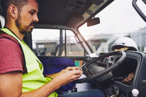 A commercial vehicle driver in a neon yellow vest sits in his truck and uses his cell phone during a rest break.