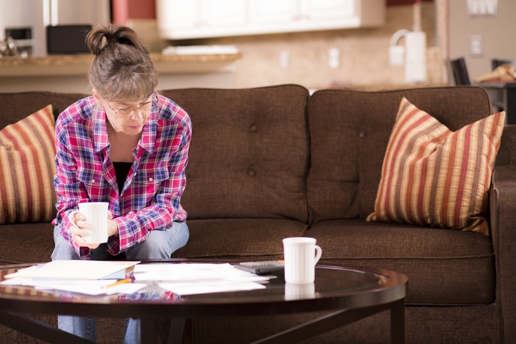 A middle aged woman in a red and blue flannel sits in her living room looking over paperwork that is spread on her coffee table.