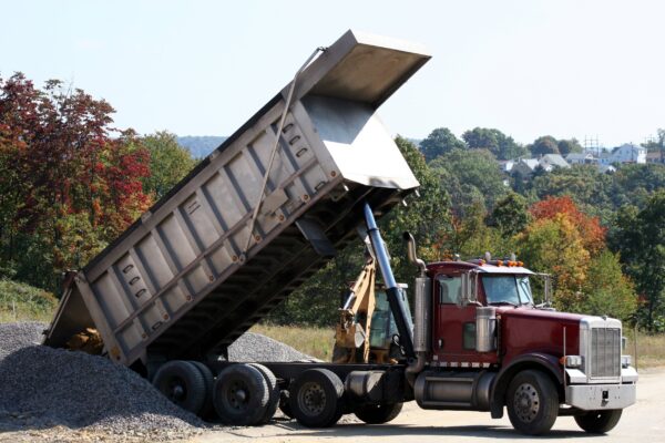 Maroon dump truck depositing gravel into a pile on the side of the road