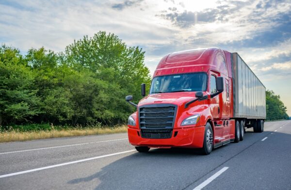 A tractor trailer truck with a bright red cab driving past lush green trees.