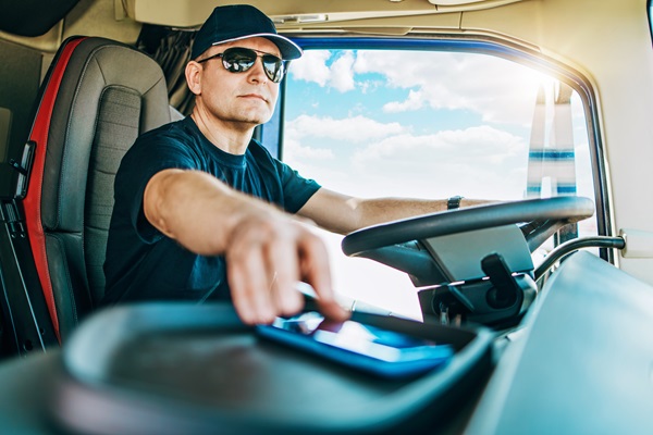 A commercial truck driver wearing sunglasses and a cap reaching for a smartphone on the dashboard while driving, illustrating a common cause of distracted driving accidents. (673159931)