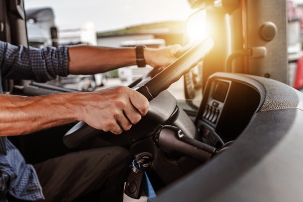 A close-up, interior view of a truck driver’s muscular arms and hands gripping a large steering wheel while driving. (195411285)