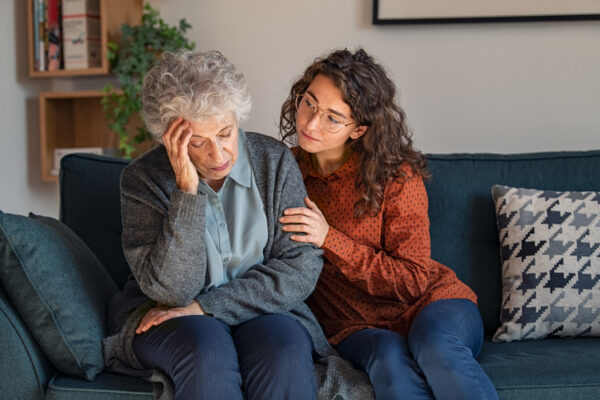 A gray-haired woman holds her head in pain on a couch as her curly-haired granddaughter comforts and supports her.