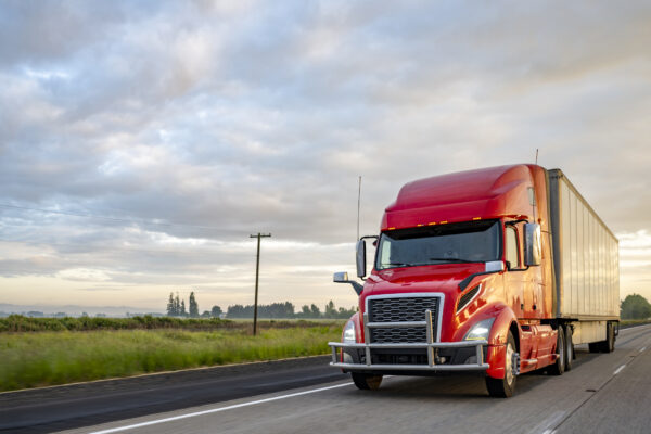 A red semi-truck cab and trailer speeding down a highway near a field.