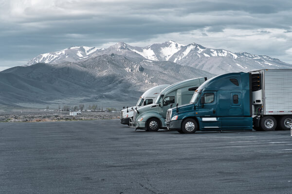 Three tractor-trailer trucks lined up at a truck stop with large, icy mountains looming in the distance.